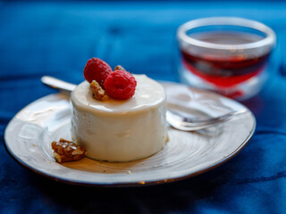 raspberry mousse cake with a glass mug of red tea and a spoon on a white plate on blue textile background, close-up side view, horizontal.