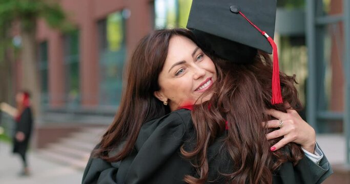 Happy mother and her student daughter on graduation day embracing with each other. Girl in academic gown and cap hugging her mother in front of the camera