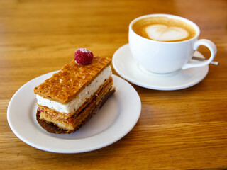 Berry milfey cake on a white plate with cup of cappuccino coffee on wooden table, horizontal, side view. Breakfast or snack or launch in cafe