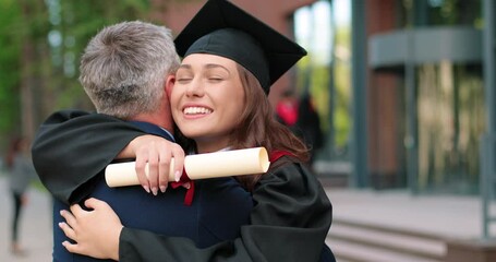 Happy female graduate are embracing with her father with diploma at her hands and rejoicing with each other. Man is hugging his daughter with other students moving and talking in background