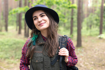 Happy young woman in a hat is smiling and holding her backpack while traveling in forest and exploring local nature 