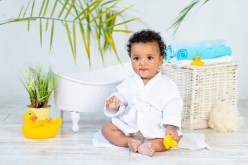 an African baby in a white coat after a bath and bathing plays with a duck at home, the concept of...