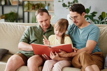 Gay parents sitting on sofa and reading a book to their little daughter in the room