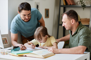 Fototapeta premium Little girl sitting at the table and doing her homework while gay parents helping her