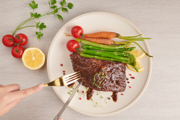 Pork roasted, grilled spare ribs from a summer BBQ served with vegetables, asparagus, baby carrots, fresh tomatoes, spices in white plate. woman hands with fork and knife eating spare ribs. top view.
