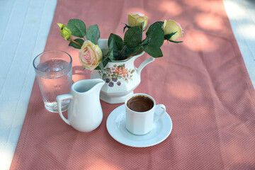 On the table in the rays of the sun there are a Turkish coffee in a white сup and a white milk jug and a glass of cold water and a vase with a bouquet of roses