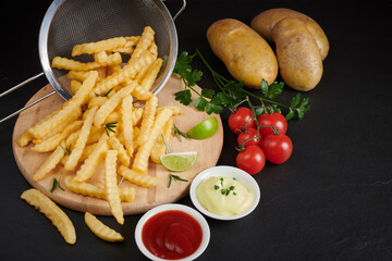 Homemade baked potato fries with mayonnaise, tomato sauce and rosemary on wooden board. tasty french fries on cutting board, in brown paper bag on black stone table background, unhealthy food.
