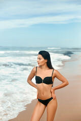 Obraz premium Portrait of young woman in black bikini on tropical beach looking at camera. Beautiful latin girl in swimwear with copy space. Sexy tanned body.