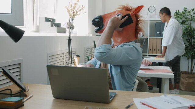 Angry businessman wearing horse mask is talking on mobile phone and gesturing sitting at desk in office while coworkers are having fun in background