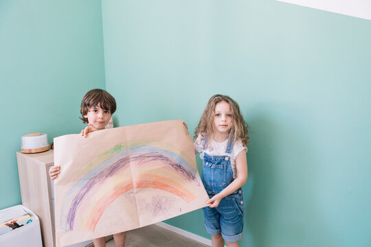 Joyful Children Decorating Wall With Rainbow Drawing And Looking At Camera