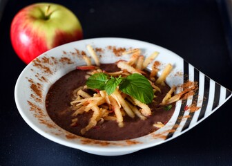 Brown soup with oatmeal, apple, milk, chocolate and mint on a white and black  plate on wooden background.