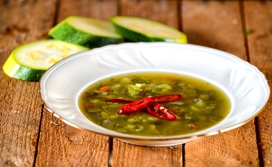 Green soup with zucchini, chilli papper and green bean on a white plate on wooden background.