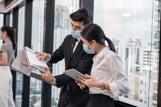 Caucasian businessman discussing and analyzing business plan with asian businesswoman holding tablet in the office. Colleagues wearing face mask protective pandemic of Covid-19