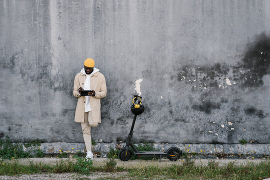 Afro man in a wall of a park using a mobile with an electric scooter parked aside