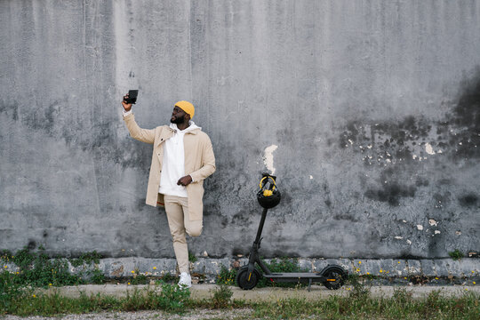 Afro man in a wall making a selfie with a mobile with an electric scooter