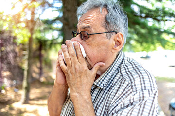 Portrait of modern senior man wearing eyeglasses allergic to the pollen blowing his nose on a summer day. Shot of an old man blowing his nose outdoors. Cropped shot of a man suffering with allergies.