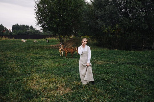 beautiful young woman in a light dress and a scarf on her head on a green meadow with goats. Agriculture.