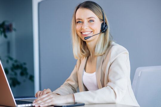 Joyful female call center operator in headset looks at the camera, smiling. Smart business woman sits at the table in the office, works in the support service, advises people