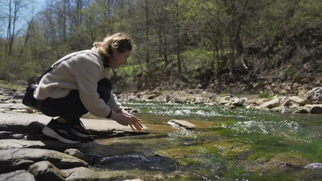 A white girl stands on a rock by the mountain river and washes her hands in the refreshing running water. The stones at the bottom of the river are covered with moss. Woman enjoys traveling.