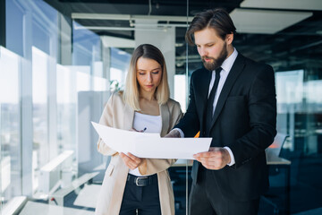 Business man and woman talking in the hallway of an office building during a coffee break. They look at the documents, discuss the contract for the deal.