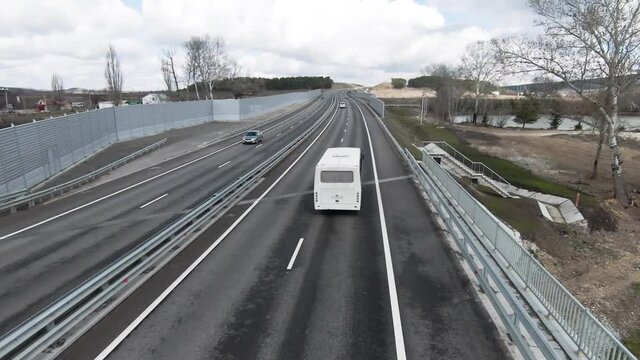 Aerial view of a white intercity bus riding through concrete straight road among steel fence, fields, and village. Countryside road with cars on a summer day.