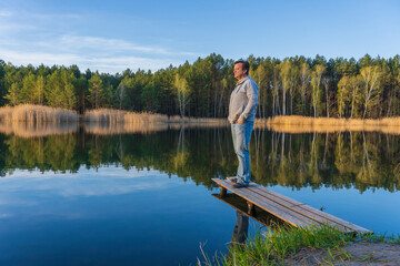 Man stands on a wooden pier near spring forest on a calm lake in Ukraine. Nature and travel concept