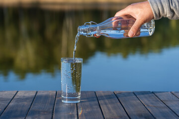 Pouring drink water into a glass from a bottle in the morning next to the lake. Nature and travel concept