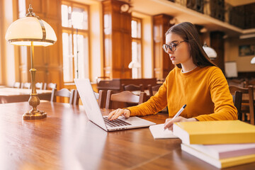 . A young student sits on a chair at a table in the library, she studies and uses a laptop to take...