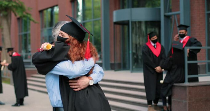 Happy father and his student daughter wearing protective mask on graduation day embracing with each other. Girl in academic gown and cap hugging her father in front of the camera
