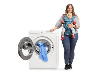 Full length portrait of a mother with a baby in a carrier standing next to a washing machine