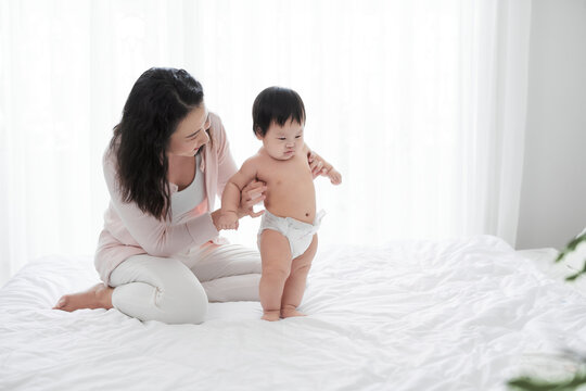 A young mother supports and teaches the baby to walk. The baby stomps on the bed.