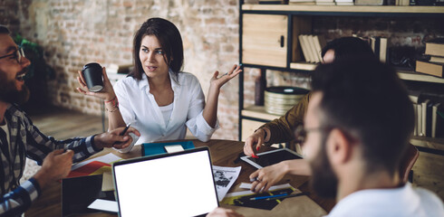 Group of young male and female coworkers collaborating on business project sitting at table with mockup laptop computer and discussing ideas, skilled hipster guys talking about startup ideas