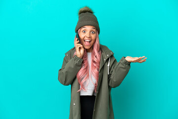 Young woman with pink hair wearing a rainproof coat isolated on blue background keeping a conversation with the mobile phone with someone