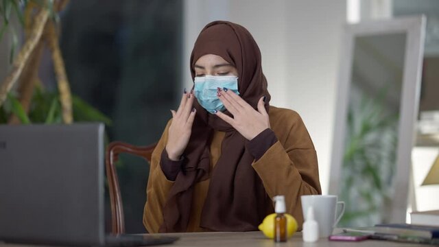 Medium shot of young Muslim woman in hijab and Covid face mask conferencing in home office sneezing showing nasal spray. Ill Middle Eastern freelancer with symptoms of coronavirus disease on isolation