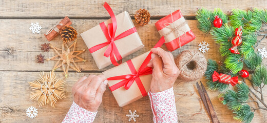 Senior woman packs gift for Christmas, top view. Wrinkled hands decorate surprise box with red ribbon, pine cones, holiday decoration, craft tools, vintage wooden table. Sharing family tradition.