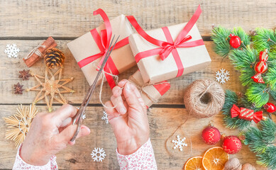 Senior woman packs gift for Christmas, top view. Wrinkled hands decorate surprise box with red ribbon, pine cones, holiday decoration, craft tools, vintage wooden table. Sharing family tradition.