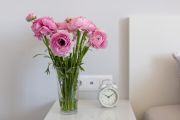 a bouquet of pink ranunculus flowers in a transparent vase on the bedside table in a bright bedroom