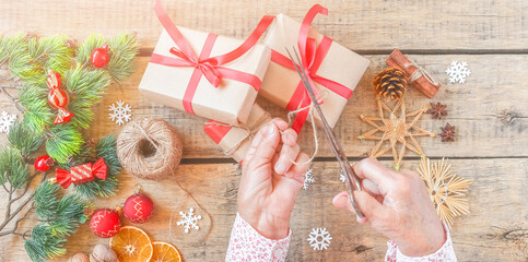 Senior woman packs gift for Christmas, top view. Wrinkled hands decorate surprise box with red ribbon, pine cones, holiday decoration, craft tools, vintage wooden table. Sharing family tradition.