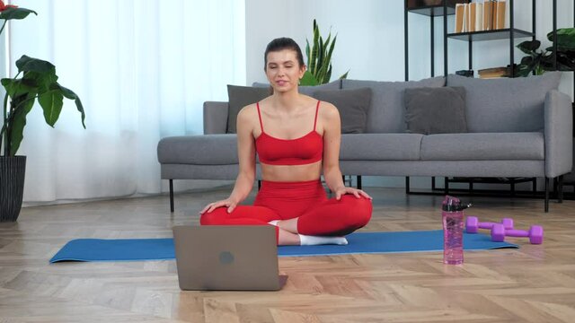 Smiling fit woman in sportswear sits on yoga mat greets waving hand talk listen fitness trainer, study online video conference call webcam chat laptop computer, watch distance sport education lesson
