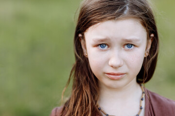 Fototapeta premium Portrait of sad crying emotional cute little girl looking at camera with face of deep sadness and sorrow outdoors. Sad child with blue eyes, copy space.