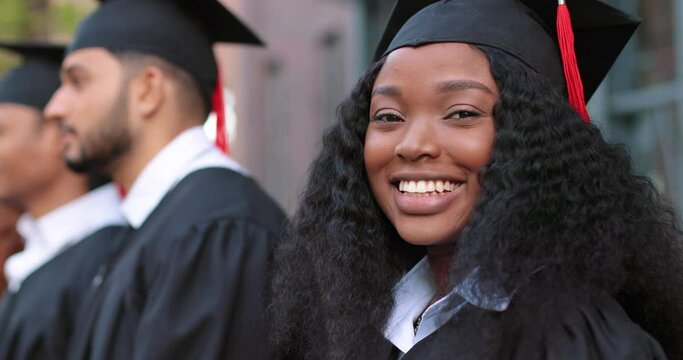 Portrait view of the multiracial happy girl looking to the camera with wide smile while standing with her group mates and listening final speech from her teacher