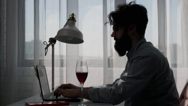 Side view of a bearded man that works at the computer full of reminder notes, man have a glass of wine near laptop to enjoy it. Home office and distance learning concept.