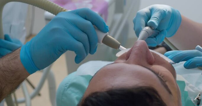 A close-up male patient wearing an optragate is having teeth cleaning by using a dental tool and saliva ejector.
