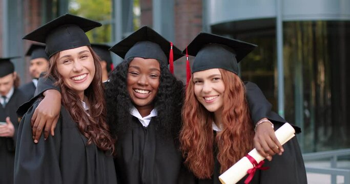 Happy group of mature students on graduation day embracing with each other. Three best girl friends in academic gowns and caps hugging in front of the camera