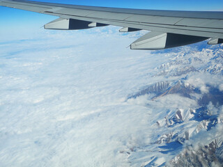 Plakat Brown snowy mountains under the clouds. View from the airplane window. Gray wing of an airplane