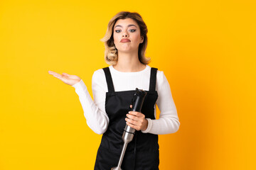 Girl using hand blender isolated on yellow background having doubts while raising hands