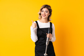 Girl using hand blender isolated on yellow background holding copyspace imaginary on the palm to insert an ad and with thumbs up