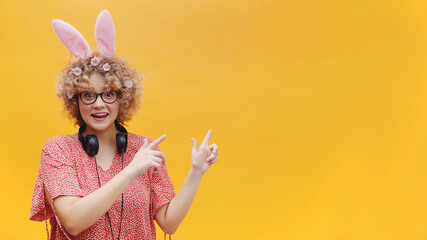 Excited young girl wearing bunny ears and glasses pointing at something. Girl in joyful mood. Headphones around her neck. Dressed in pink shirt. Isolated over yellow background studio. 