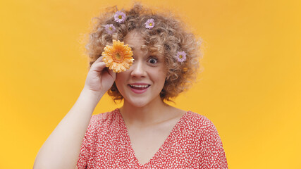 Beautiful young girl covering her eye with an orange gerbera daisy flower. Isolated girl with bright yellow background studio. Funky girl smiling and wearing flowers in her curly hair. 