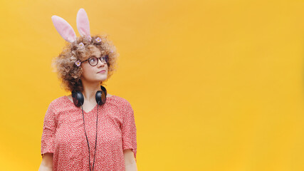 Beautiful young girl wearing bunny ears and spectacles looking away from the camera. Headphones around her neck. Dressed in pink shirt. Isolated over yellow background studio. 